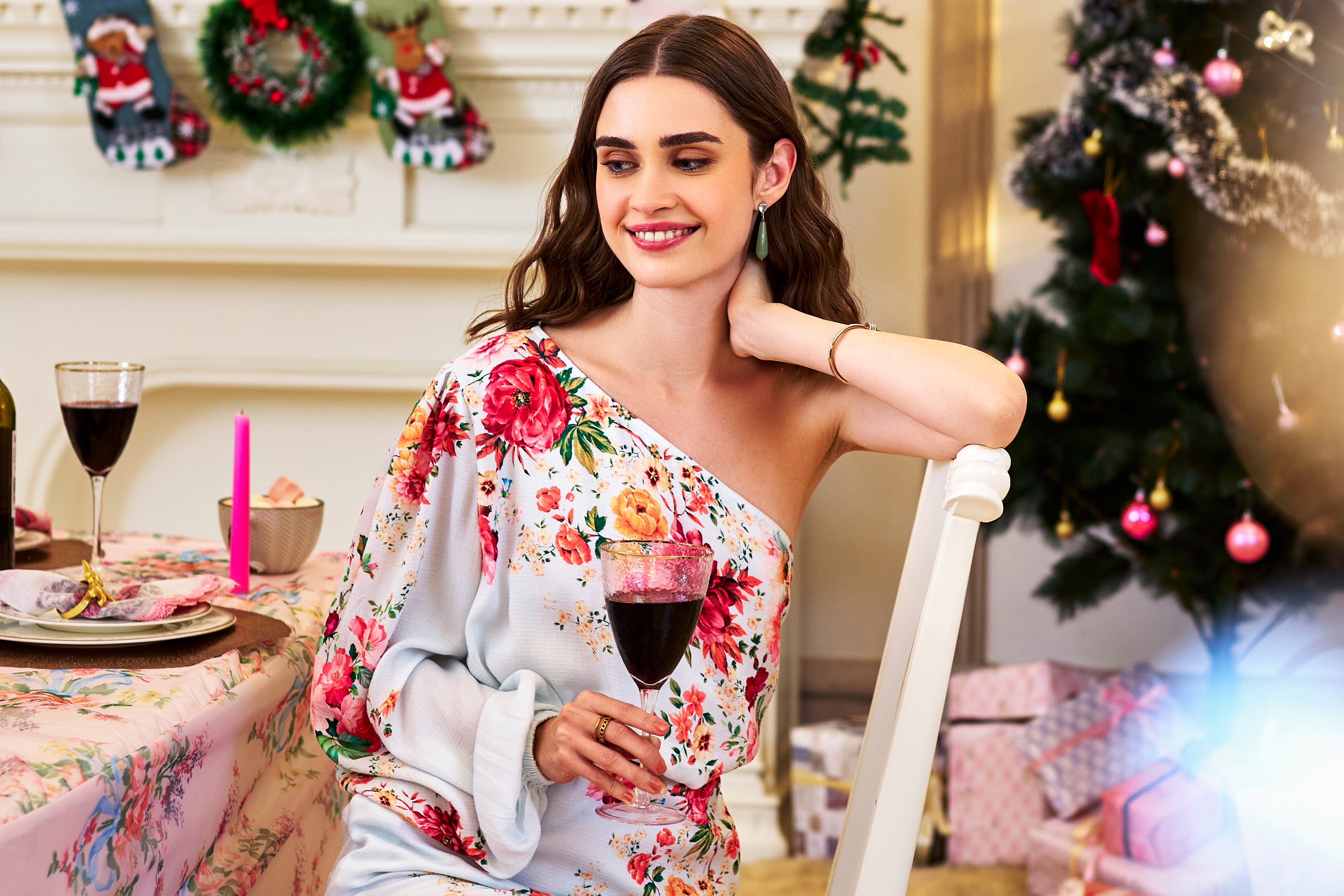 Smiling woman in a white floral one-shoulder dress, holding a wine glass, celebrating near a Christmas tree and festive table setup – Pretty Pixie holiday collection