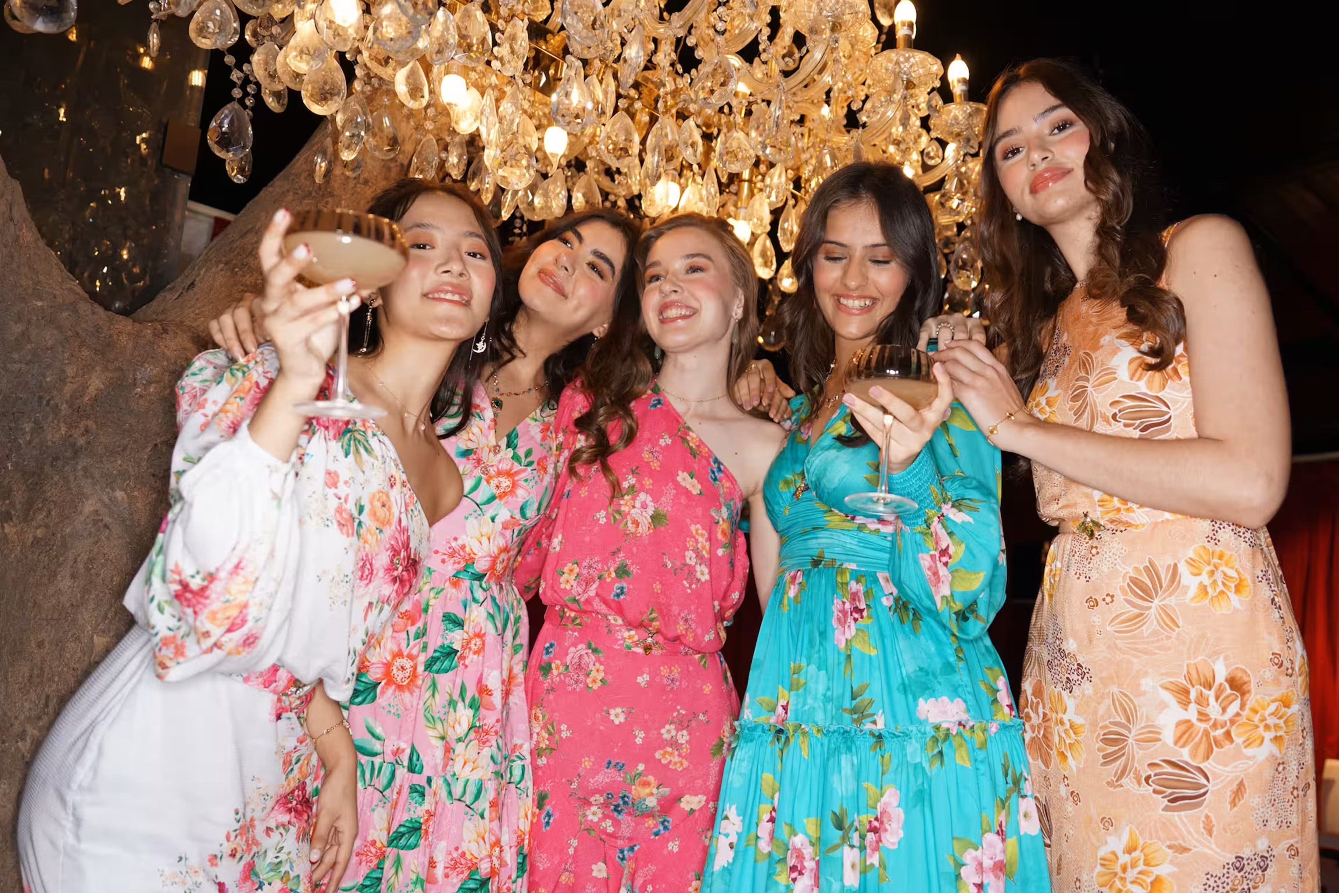 Group of women wearing colorful floral dresses at a party under a chandelier, styled for a fashion lookbook