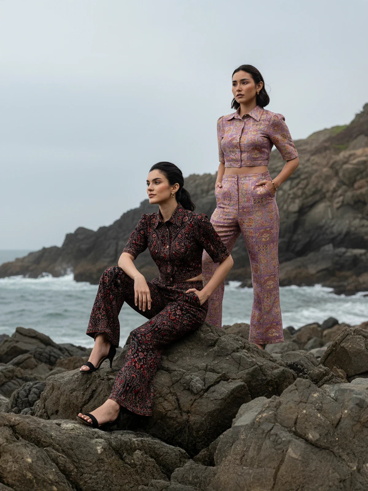 Two women wearing Pretty Pixie luxury printed co-ord sets posing on coastal rocks during an outdoor fashion photoshoot with waves in the background.