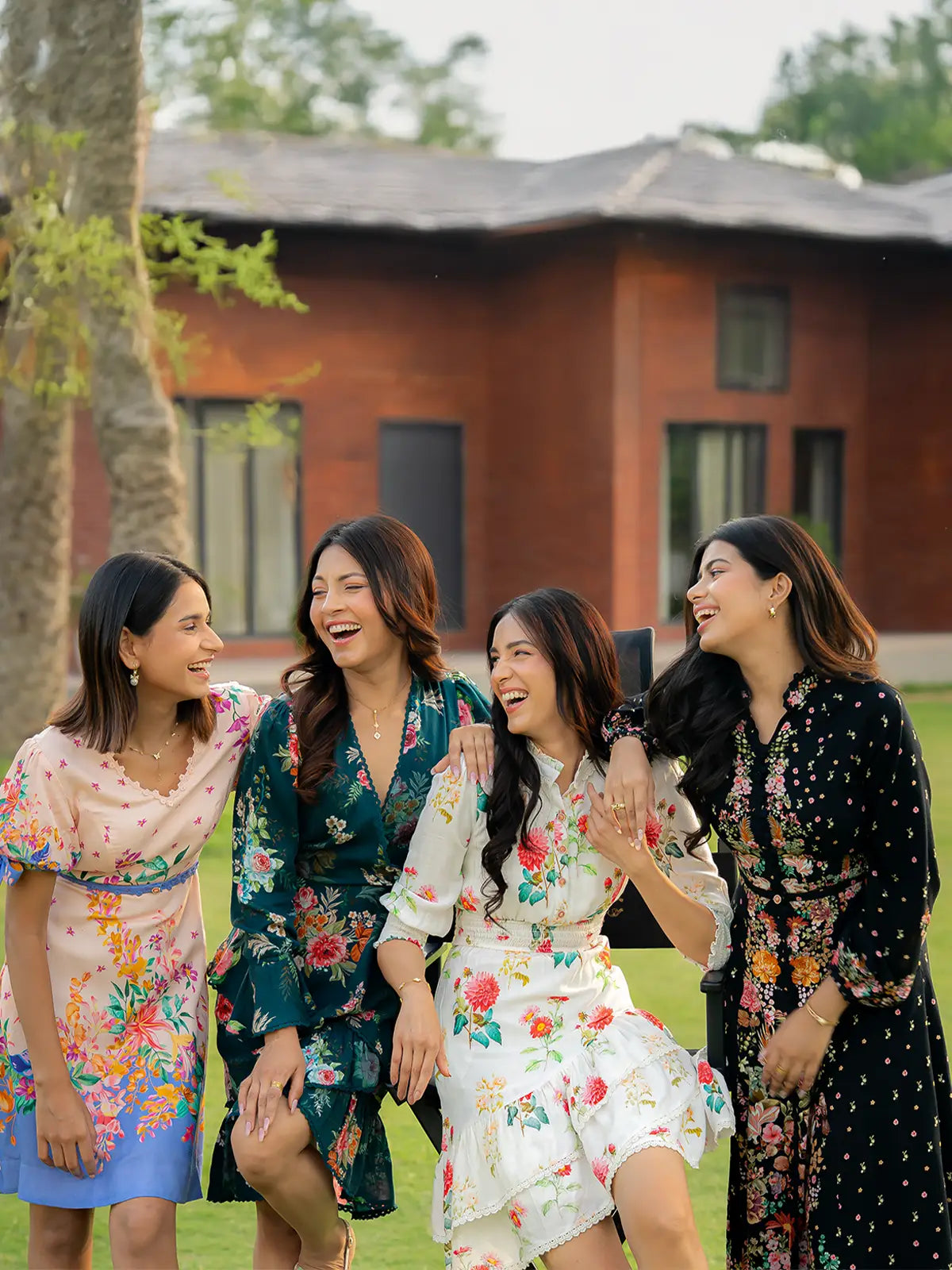 Four women wearing Pretty Pixie floral dresses in pastel and dark prints, laughing and walking together outdoors in a garden setting during a spring–summer fashion photoshoot.