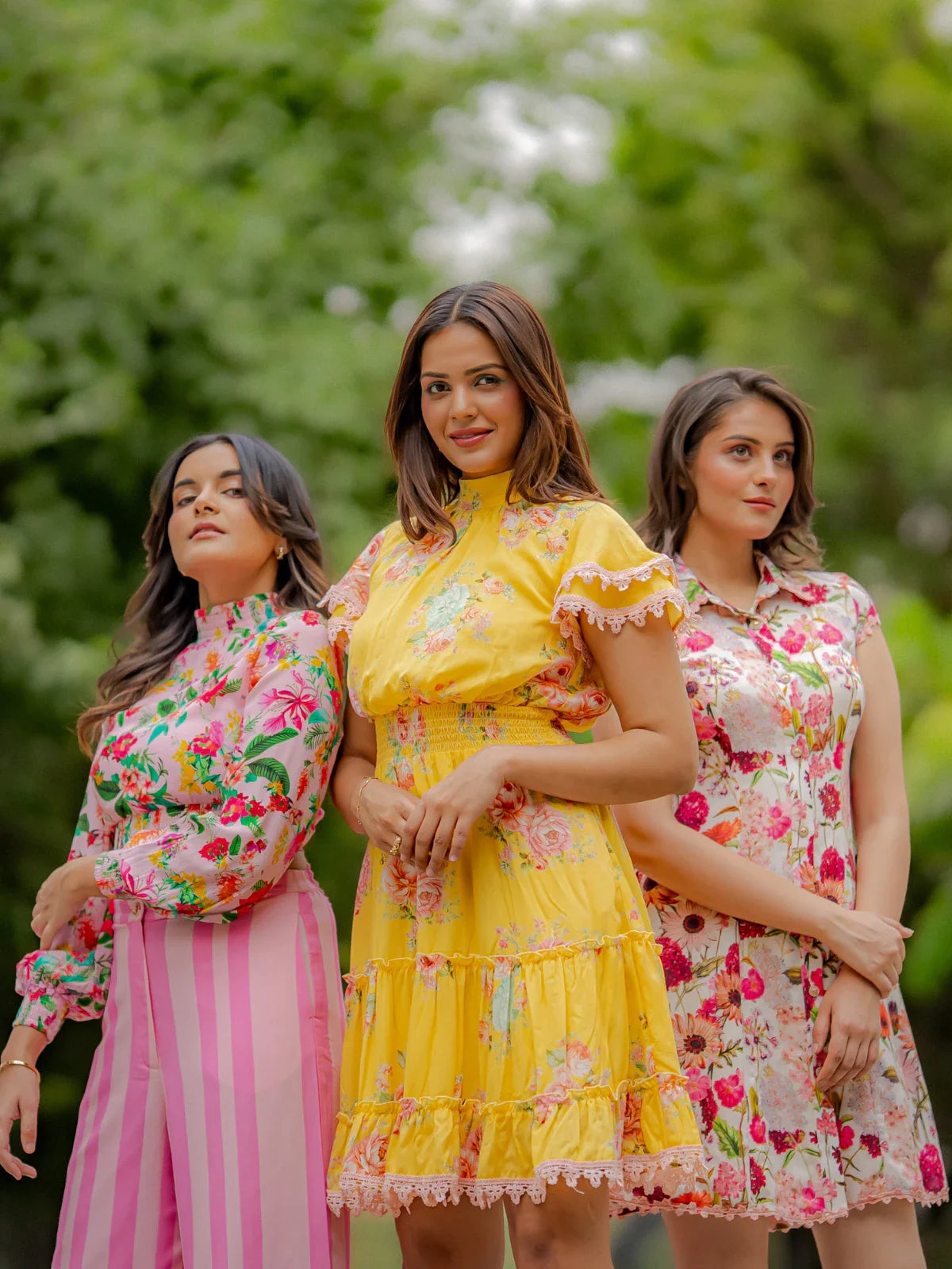 Three women wearing Pretty Pixie bright floral summer outfits—including a yellow tiered dress, pink striped pants with floral top, and a floral mini dress—posing outdoors in a lush green setting.