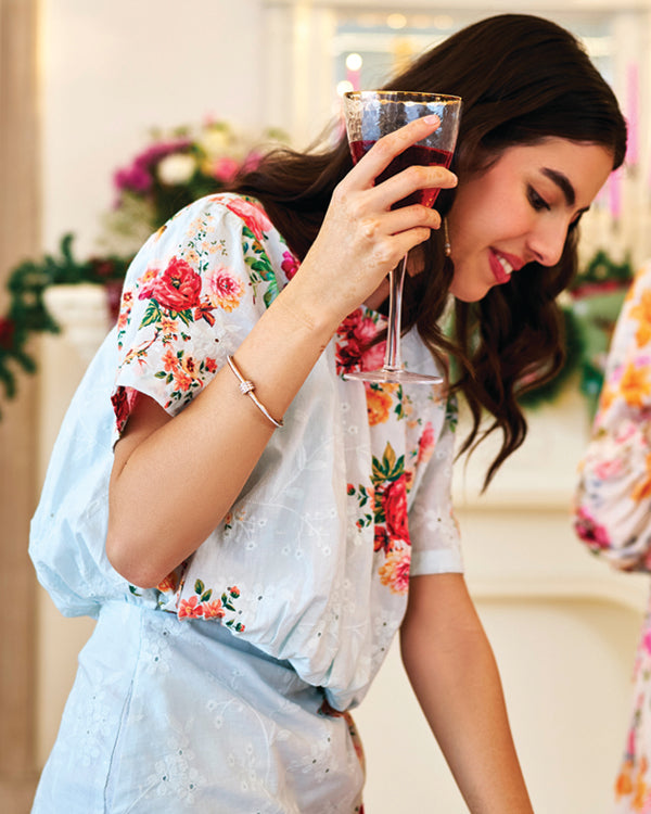 Smiling woman holding a glass, dressed in a pastel floral dress with pink and red roses – Pretty Pixie Spring Collection