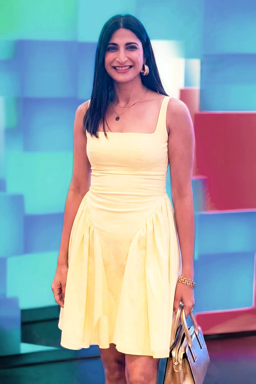Woman wearing sleeveless yellow square neck mini dress holding metallic handbag, posing against colorful geometric backdrop, Ahana style India.