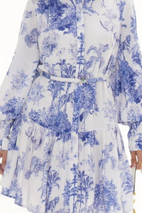 Woman in Pretty Pixie blue and white toile jungle-print belted tiered dress with butterfly brooch