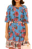 A woman elegantly poses in a blue floral dress adorned with delicate pink flowers, exuding grace and style.