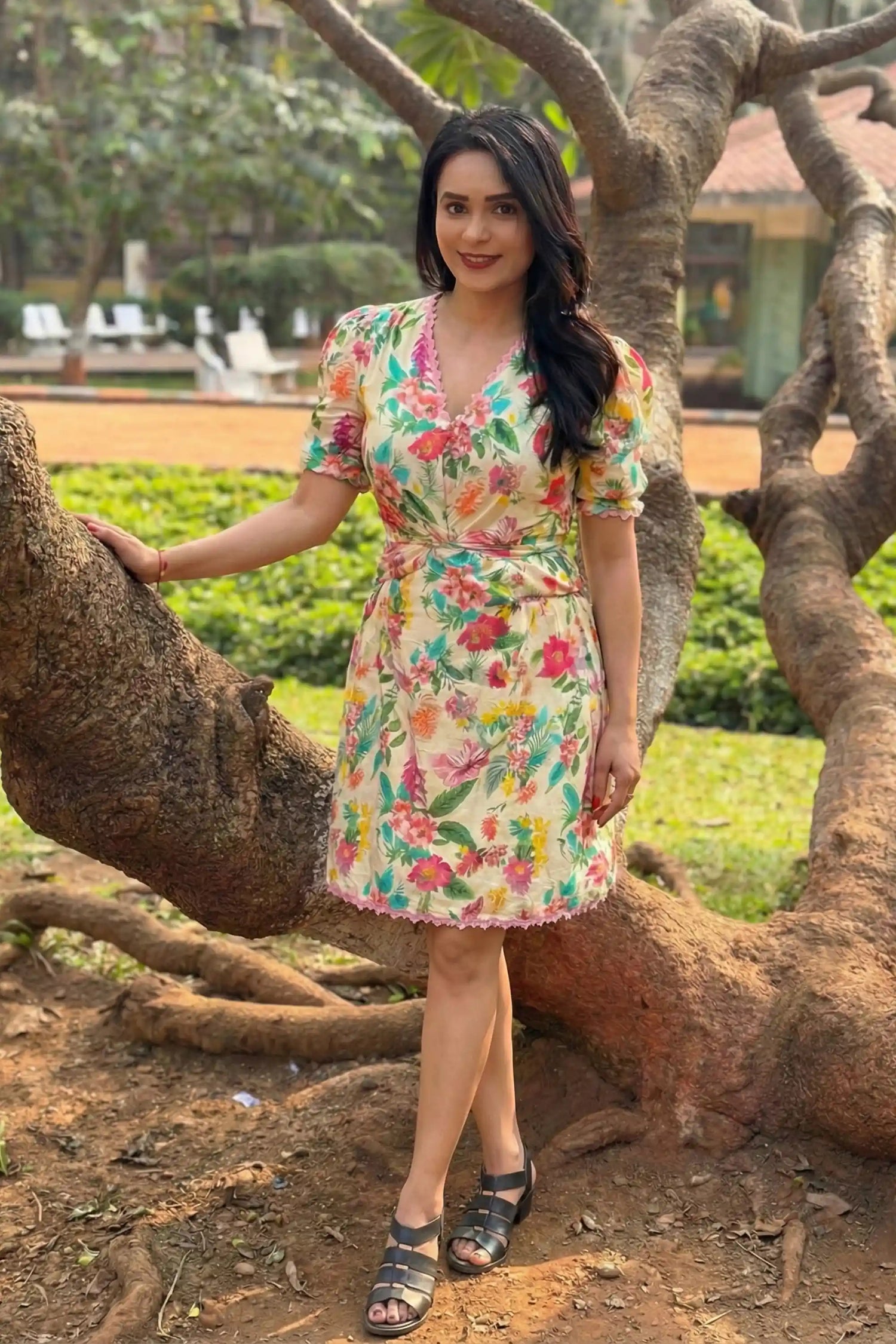 Woman wearing a floral summer dress standing outdoors near a tree in a garden setting during a natural light fashion photoshoot.
