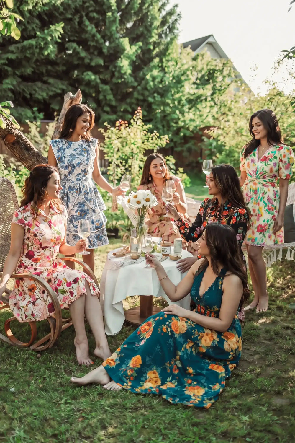 Multiple women wearing different organic cotton floral print dresses at garden tea party; mix of blue floral, pink botanical, and multicolor printed dresses