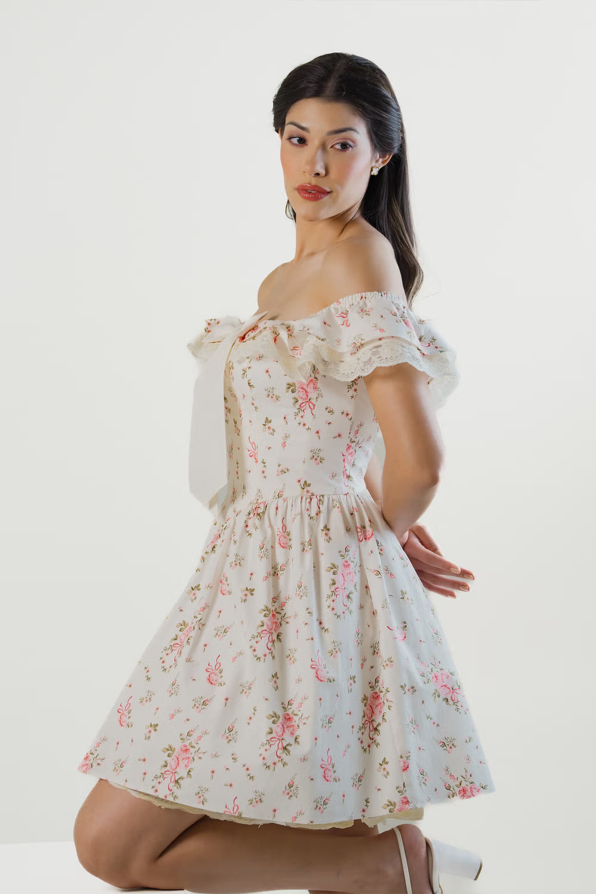 Woman posing in ivory floral mini dress with off-shoulder lace trim and statement bow.