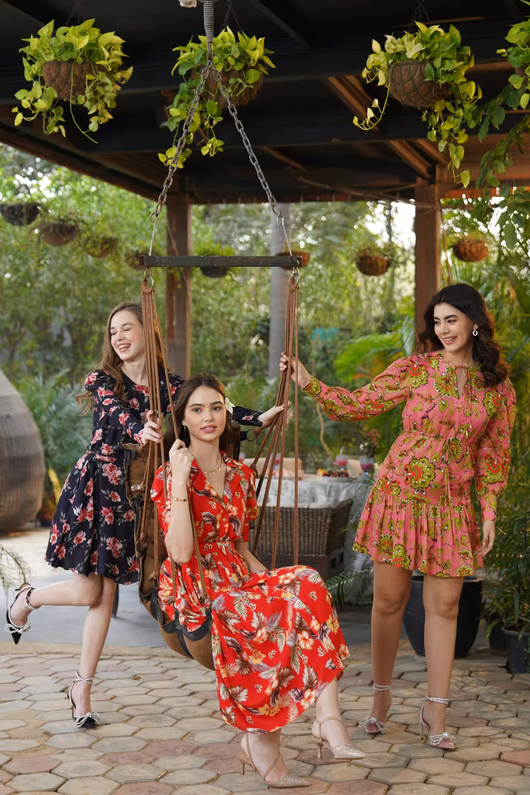 Three women in floral dresses posing together outdoors with a swing and greenery.