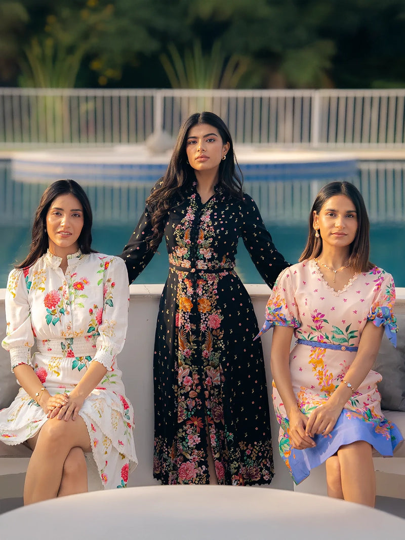 Three women in floral midi dresses sitting and standing beside swimming pool, dressed in white, black, and pastel prints, summer evening India.