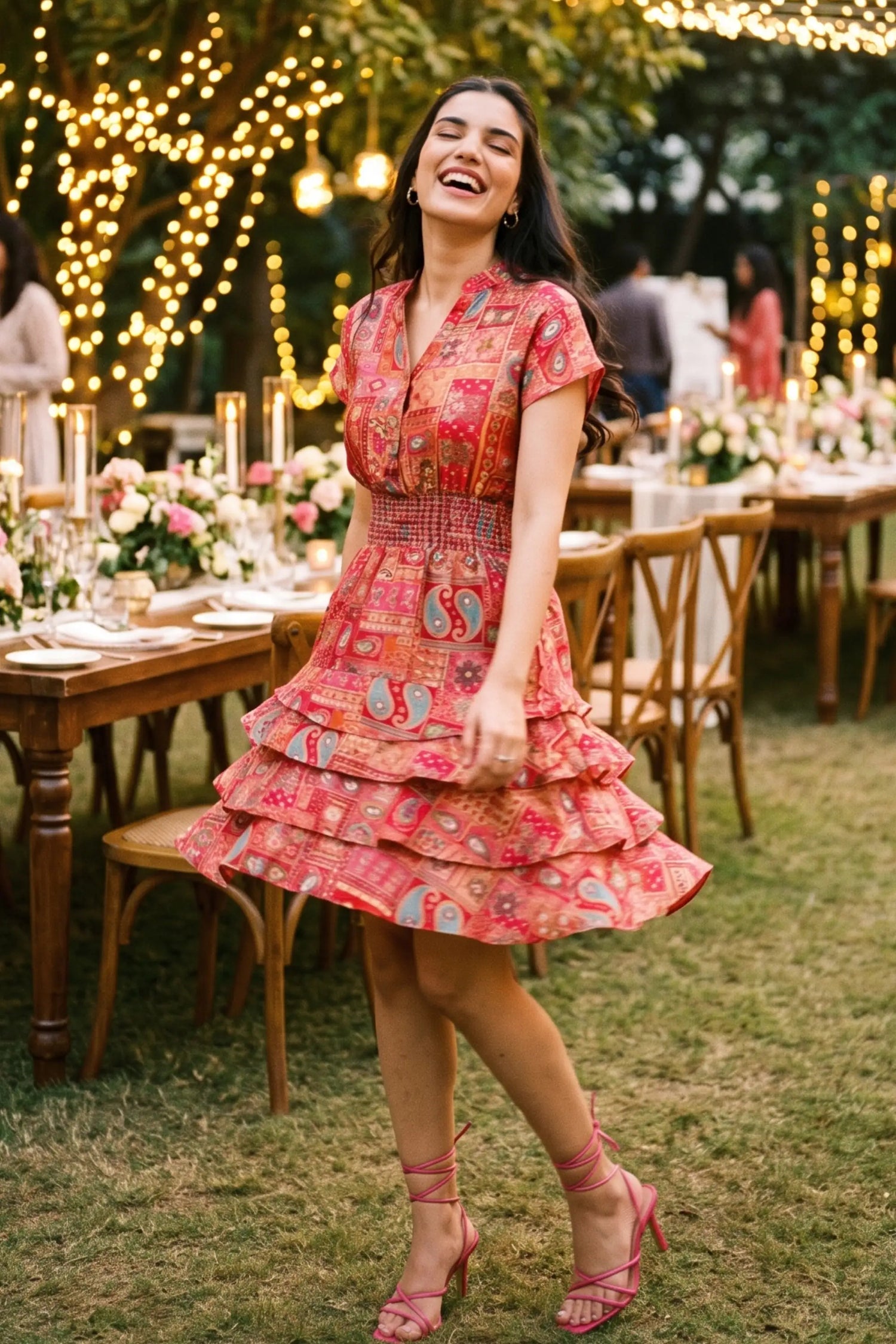 Woman twirling in coral boho red smocked waist tiered ruffle mini dress with pink strappy heels at outdoor garden party with string lights - Pretty Pixie
