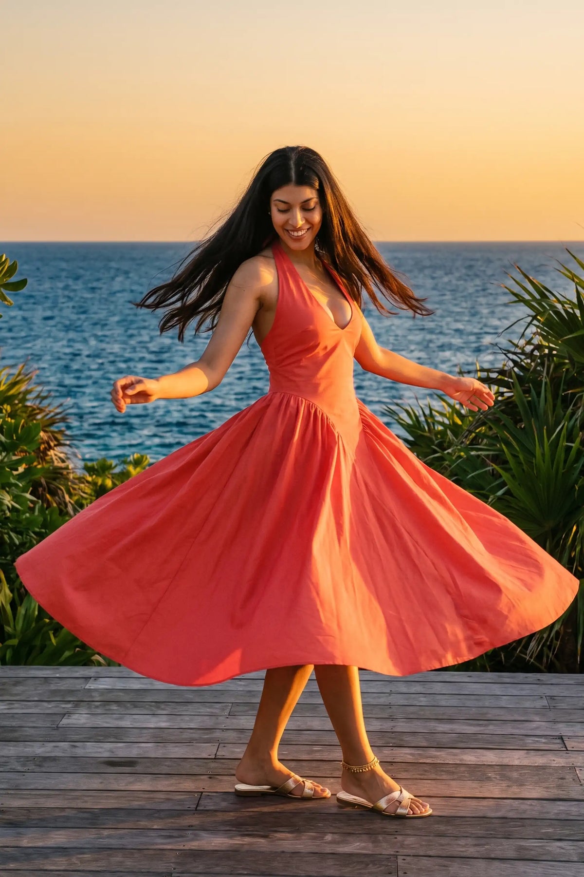 Woman twirling on a seaside deck at sunset wearing Pretty Pixie's coral red halter bustier midi dress