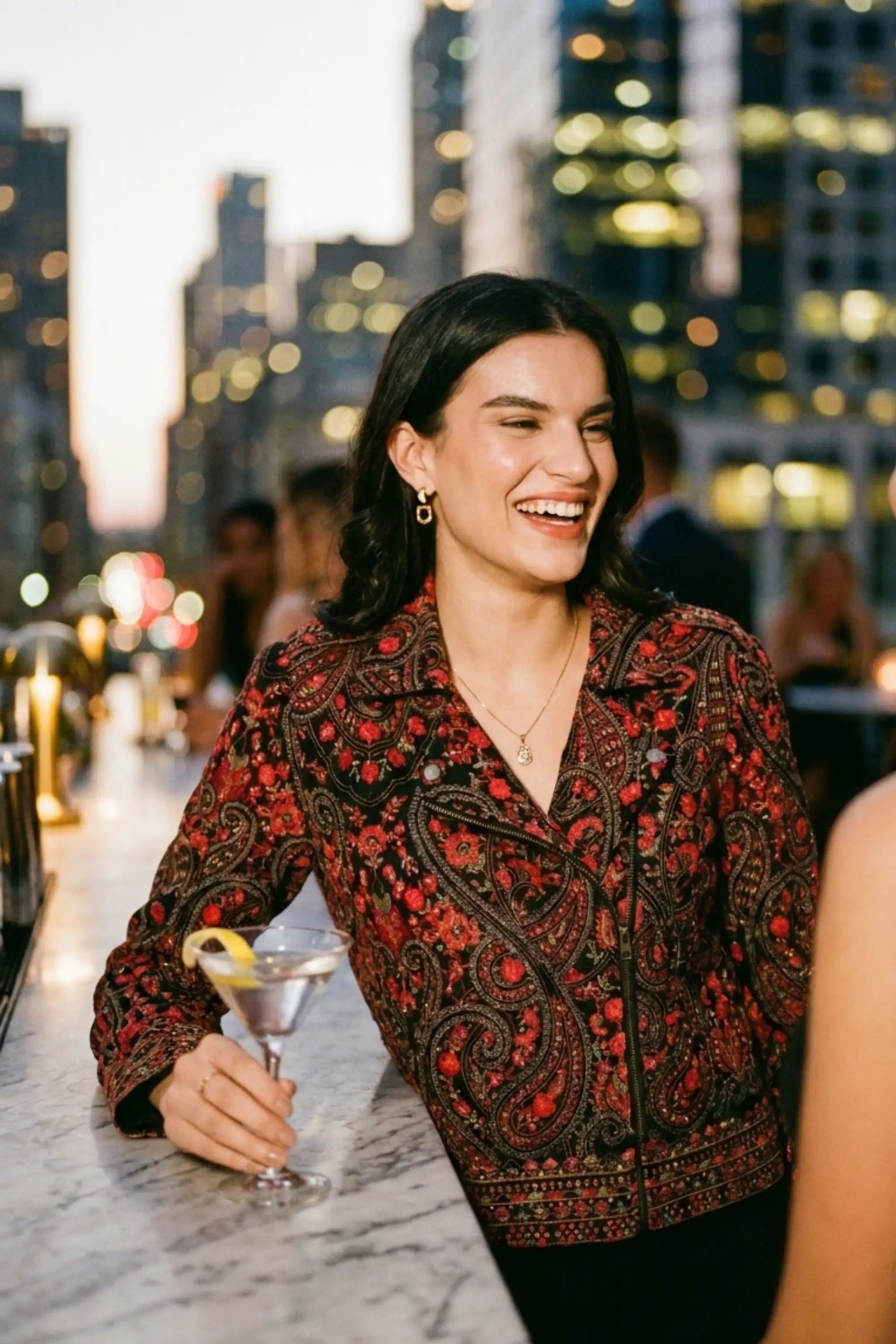 Woman in crimson paisley black hand embroidered biker jacket with gold drop earrings holding cocktail at rooftop bar with city skyline - Pretty Pixie eveningwear