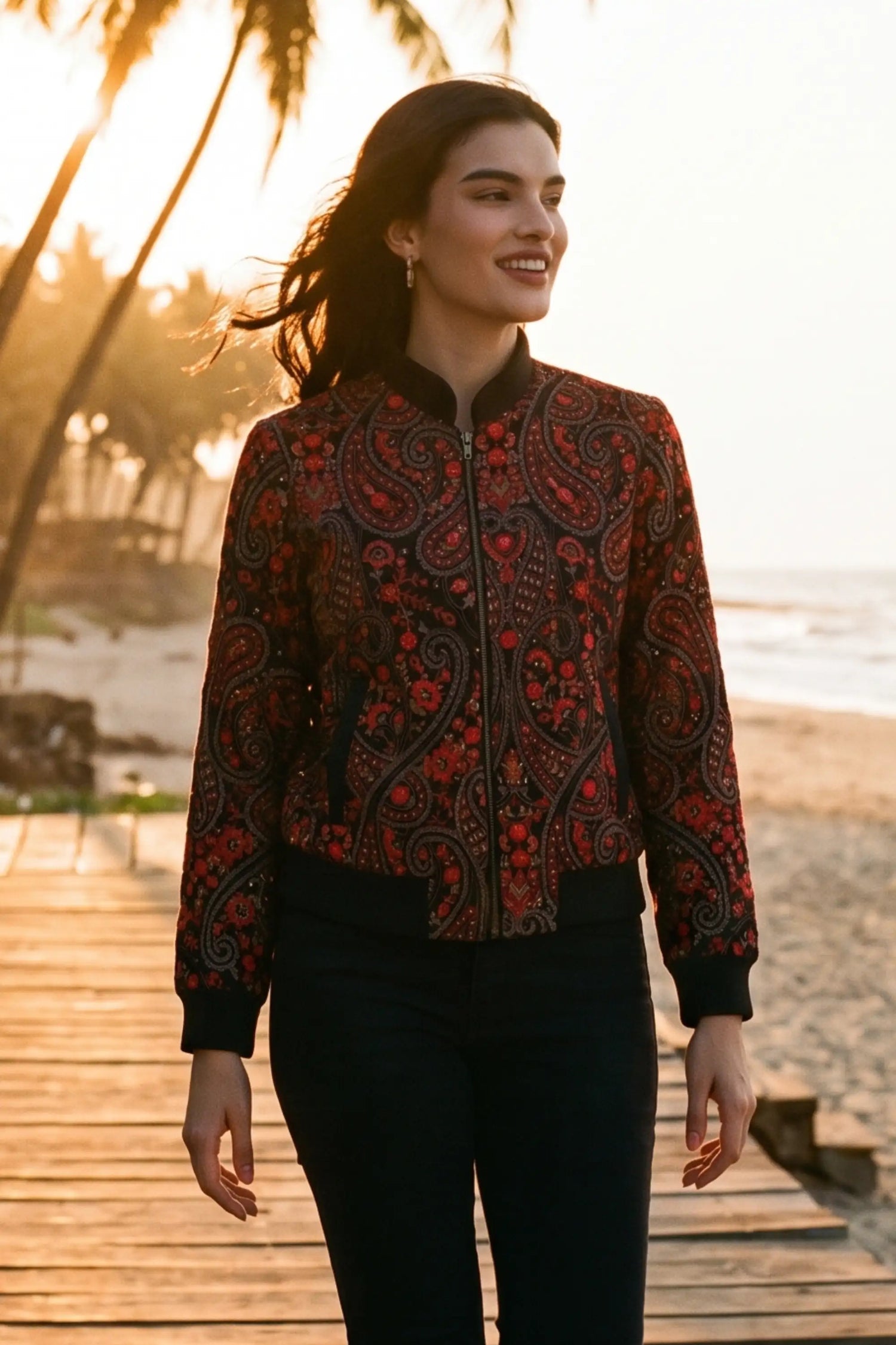 Woman in crimson paisley black hand embroidered bomber jacket with gold drop earrings walking beach boardwalk at sunset - Pretty Pixie eveningwear
