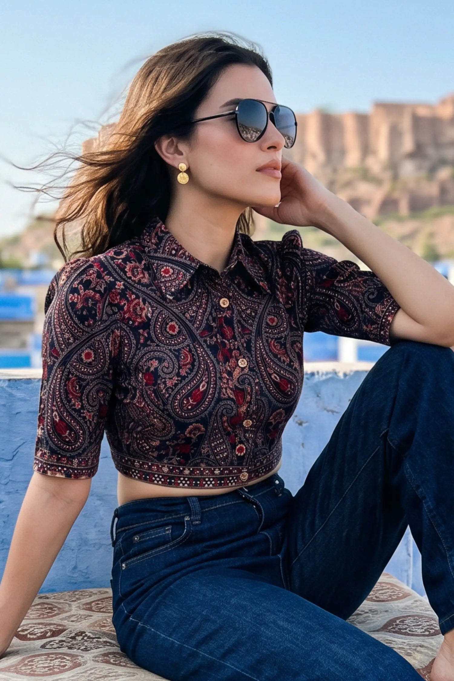 Woman in crimson paisley navy hand embroidered crop shirt with gold drop earrings and denim jeans on rooftop overlooking Jodhpur fort - Pretty Pixie travel wear