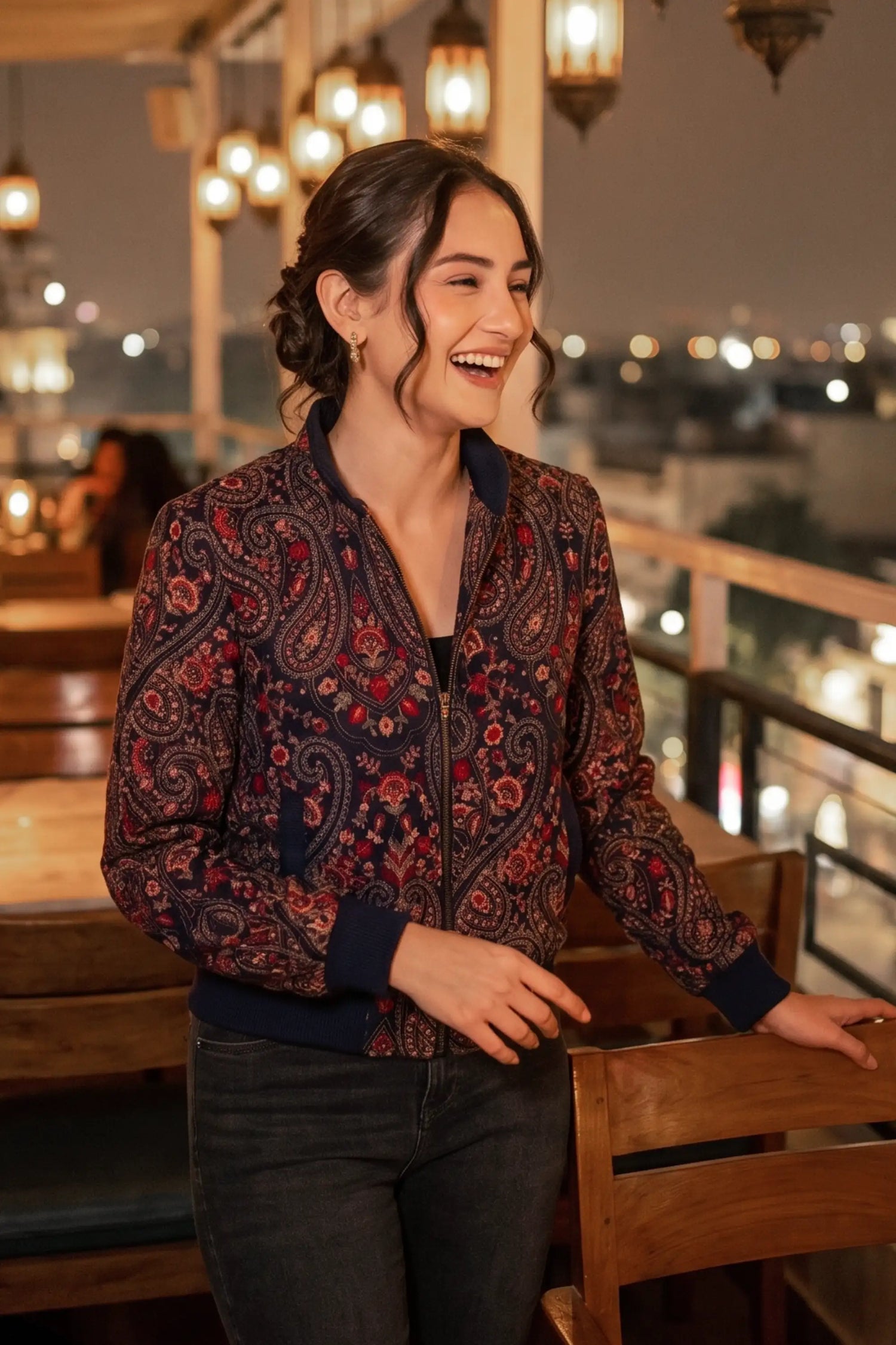 Woman in crimson paisley navy hand embroidered bomber jacket with black jeans laughing at rooftop restaurant with string lanterns - Pretty Pixie eveningwear