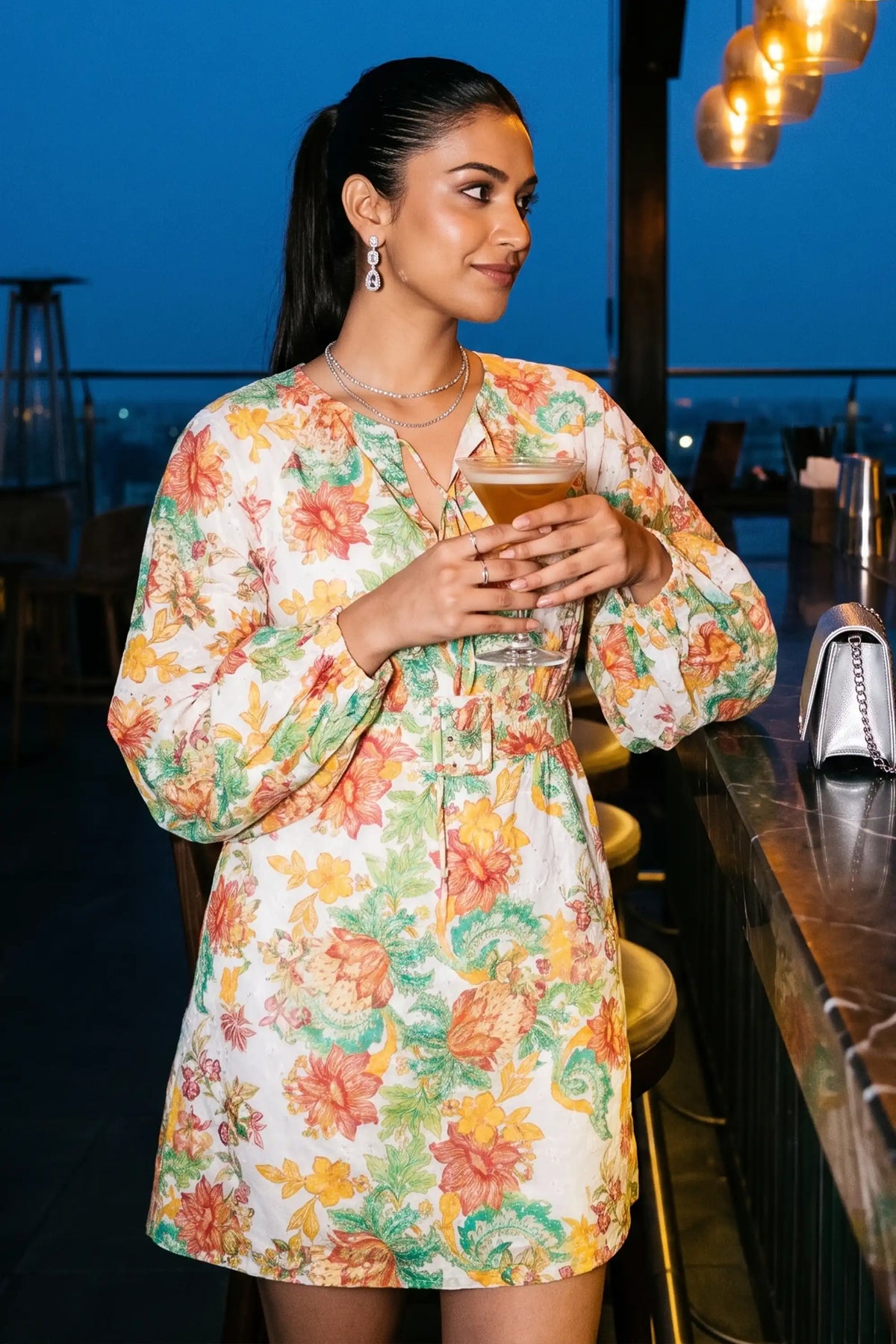 Woman holding a cocktail at a rooftop bar wearing Pretty Pixie's gardenia floral puff-sleeve smocked mini dress