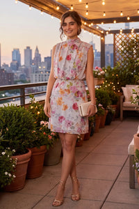 Woman smiling on a rooftop terrace with a city skyline backdrop wearing Pretty Pixie's ivory floral high-neck sleeveless mini dress