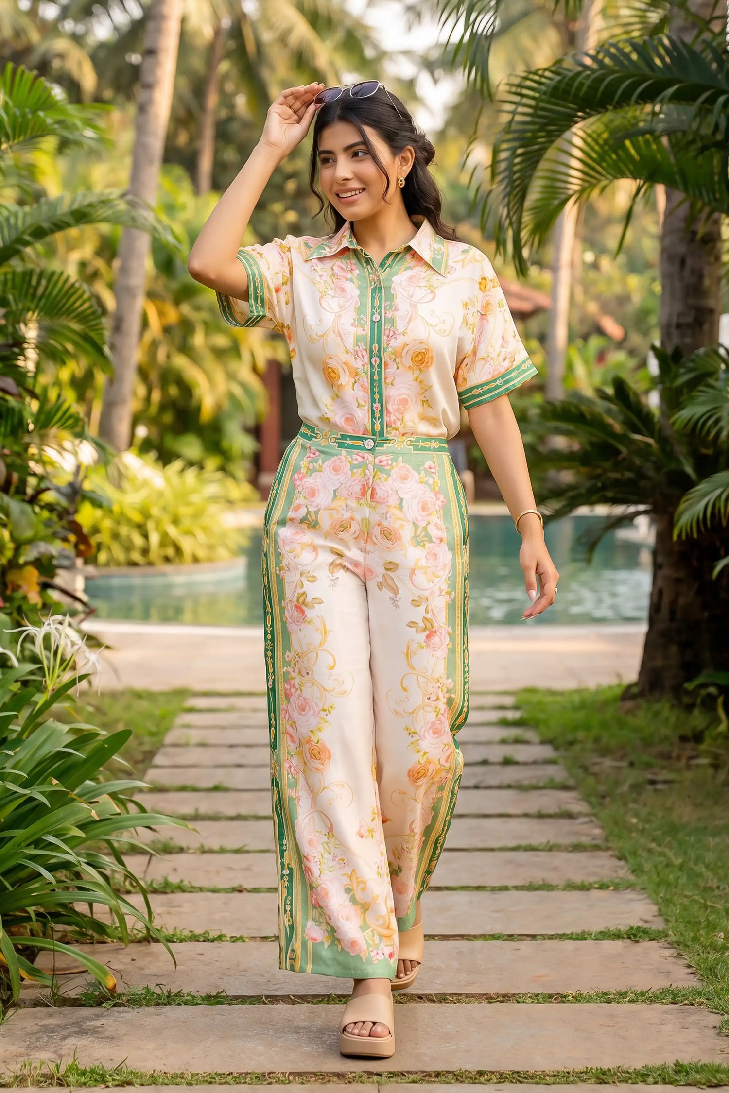 Woman in ivory and green floral collared co-ord set with striped border print walking poolside path lined with palms - Pretty Pixie resort co-ord