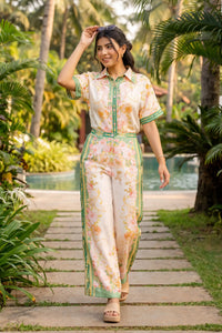 Woman in ivory and green floral collared co-ord set with striped border print walking poolside path lined with palms - Pretty Pixie resort co-ord