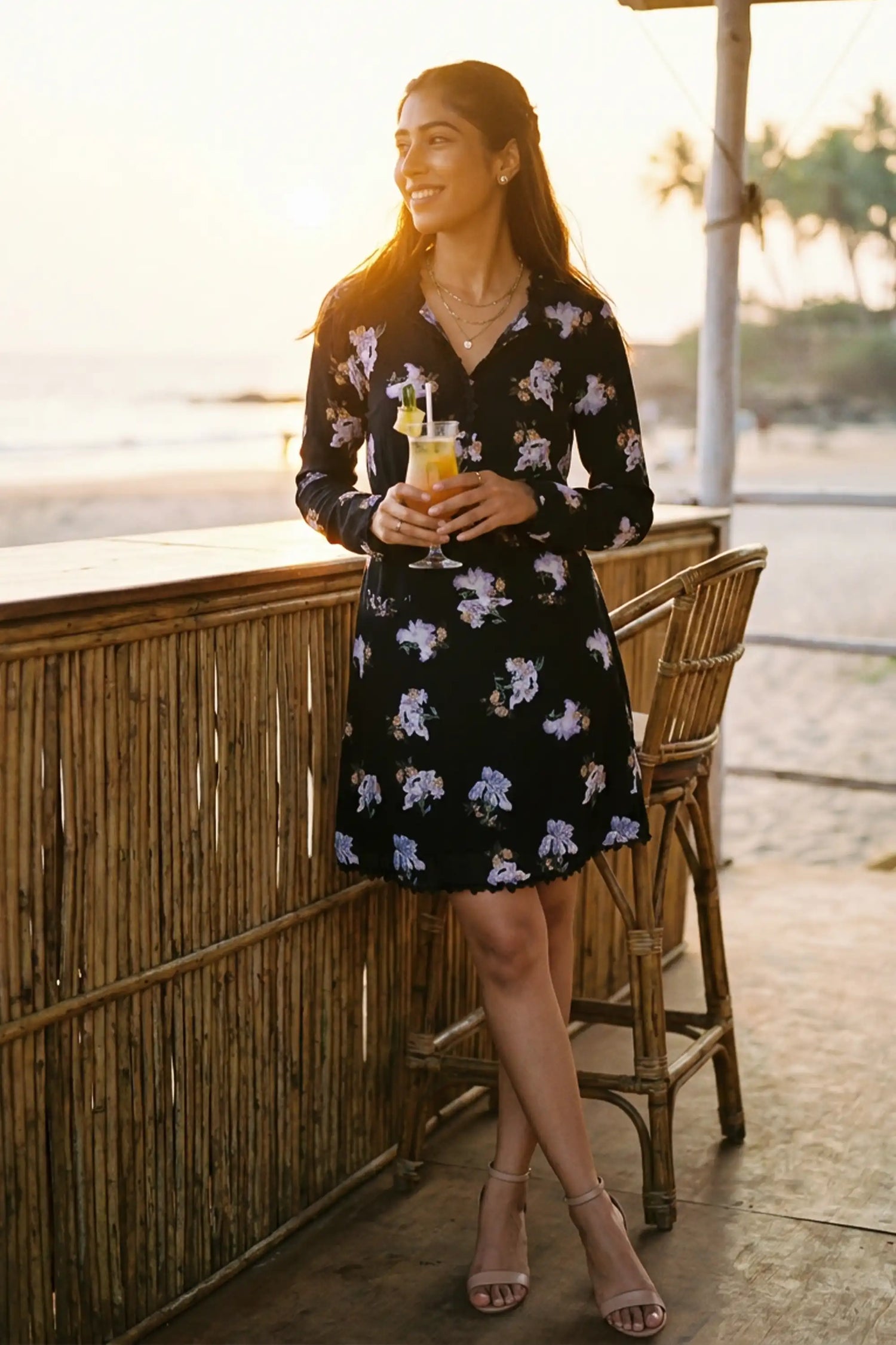 Woman in midnight iris floral belted long-sleeve mini dress holding cocktail at bamboo beach bar during sunset - Pretty Pixie resort wear