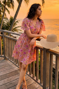 Woman leaning on a railing at beach sunset wearing Pretty Pixie's peach blossom floral V-neck flutter sleeve mini dress