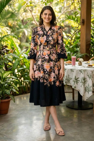 Woman in Rosalia Bloom black floral belted midi dress with bell sleeves and mandarin collar smiling on garden patio with breakfast table - Pretty Pixie daywear