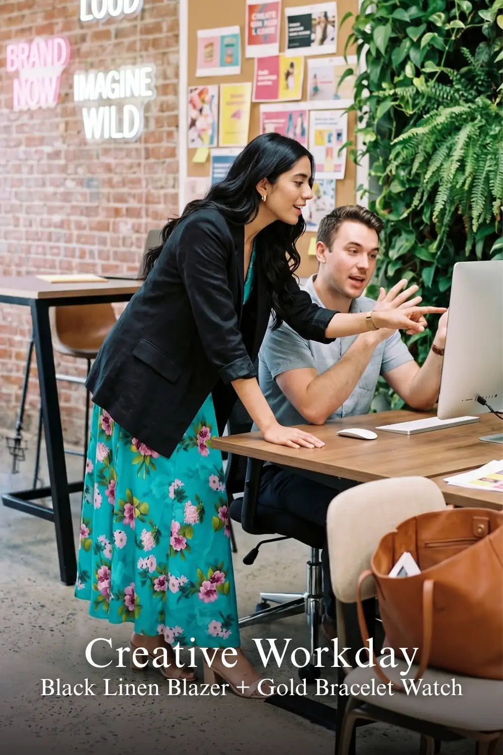 Woman in turquoise floral dress layered with black linen blazer and gold bracelet watch, pointing at computer screen in creative office