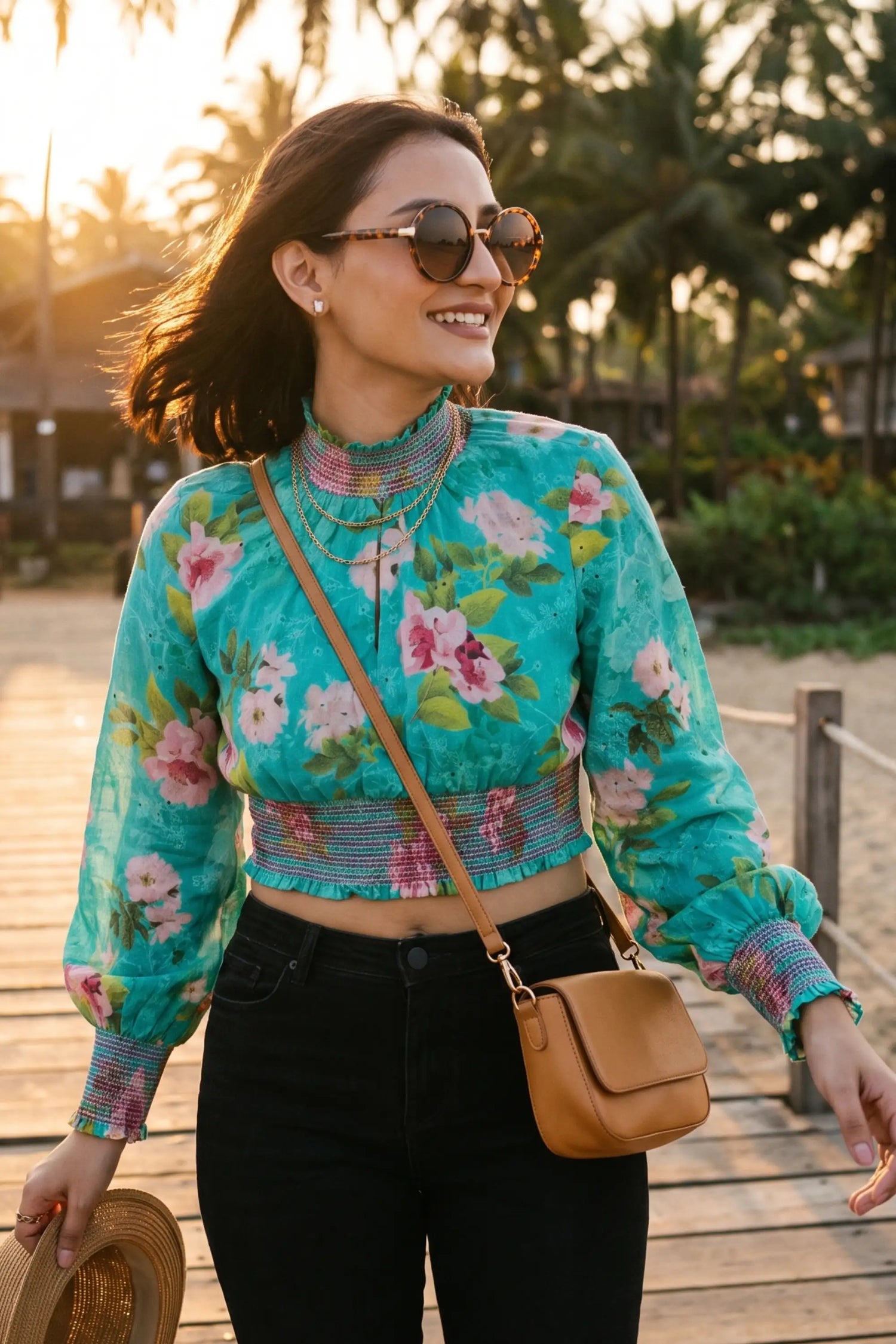 Woman in turquoise floral smocked crop top with gold layered necklace and tan crossbody bag walking beach boardwalk at sunset - Pretty Pixie resort wear