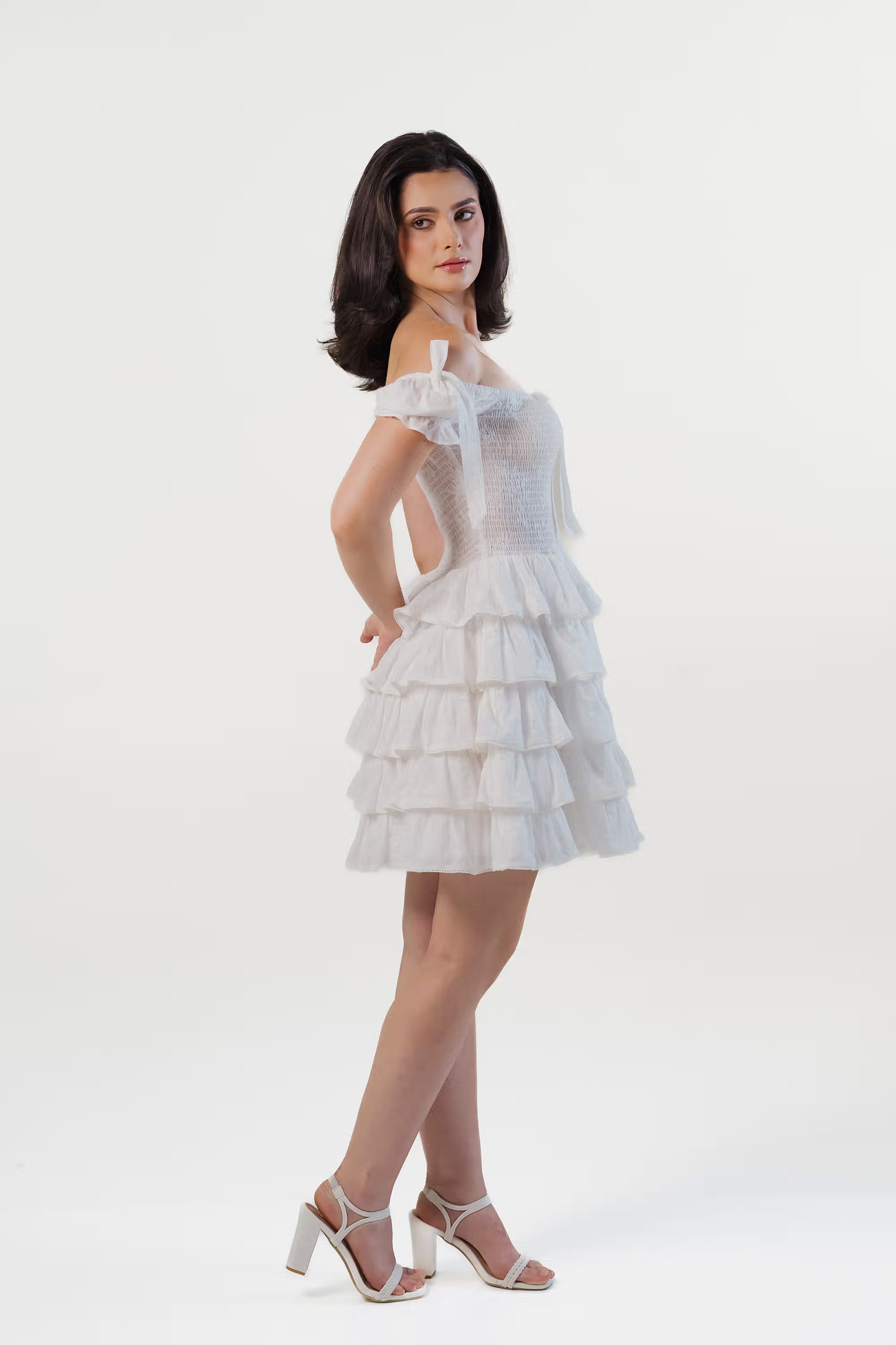 Side profile of a woman wearing a white ruffle mini dress with off-shoulder design and layered hem.